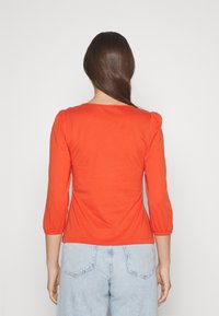 Red, fitted top with three-quarter sleeves and puffed shoulders. Made of a smooth, stretchy cotton blend. Back view, paired with light denim.