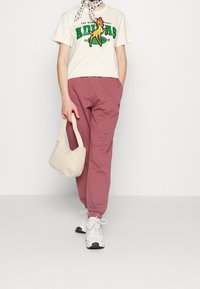 Lyhythihainen kerma-vaaleanpunainen t-paita, jossa on vihreä ja keltainen graafinen logo, yhdistetty vaaleanpunaisiin joggereihin ja karkean tekstuurin beigelle laukulle. Valkoiset lenkkarit.