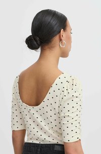 Top crema a pois con scollatura bassa, tessuto texturizzato e maniche corte, abbinato a una fascia in vita nera. Orecchino a cerchio in argento tondo.