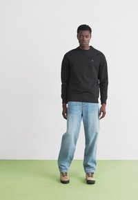Felpa nera con scollo rotondo e logo discreto, abbinata a jeans in denim blu medio. Modello in piedi in un ambiente neutro su un pavimento verde.