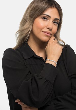 Bracciale d'argento con perle tonde indossato su un polso, caratterizzato da una superficie liscia e leggero luccichio, abbinato a una blusa nera.