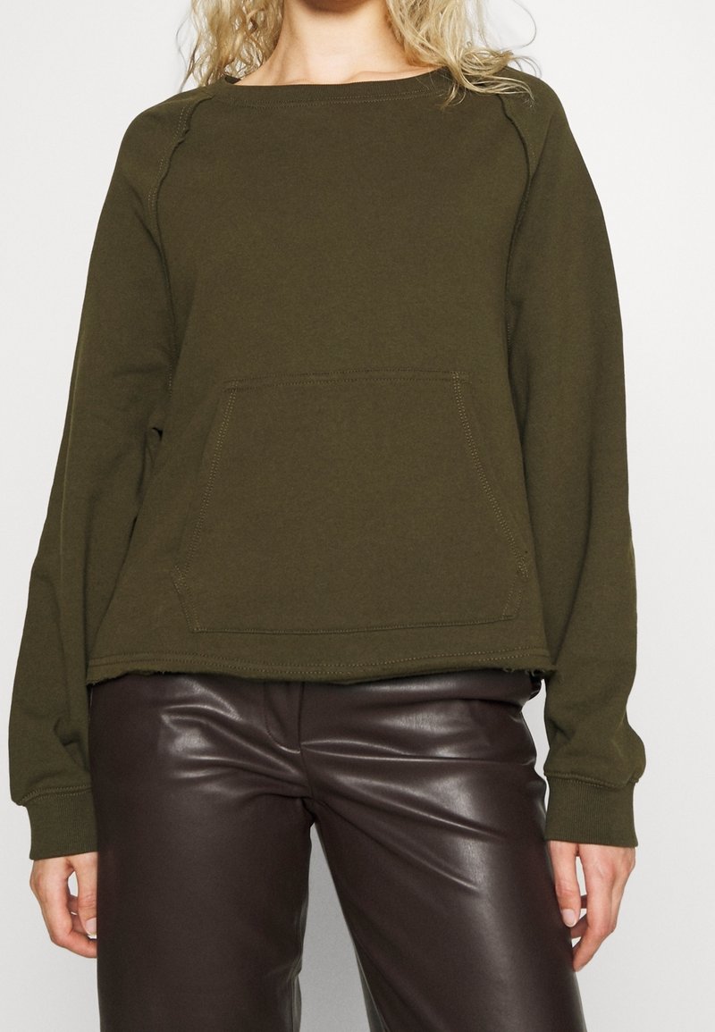 Sudadera verde oliva con escote ancho y manga larga, que presenta un bolsillo frontal y dobladillo sin rematar, combinada con pantalones de cuero marrón.