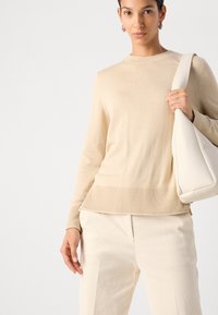 Beiger Rippenpullover mit Rundhalsausschnitt, kombiniert mit hellen Cordhosen, gehalten mit einer cremefarbenen Schultertasche.