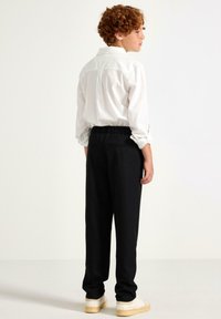 Hvid skjorte med krave og sorte elastiske taljebukser, lavet af glat stof med tapered ben, parret med beige sko. Bagfra.