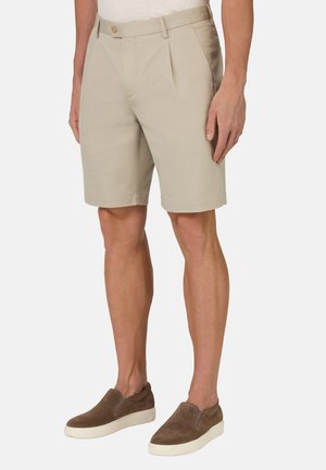 Shorts beige avec plis fronts, passants de ceinture et fermeture à bouton. Portés avec des chaussures marron à enfiler avec une semelle blanche.