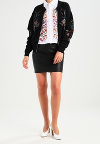 Cardigan nero ricamato sopra una blusa bianca ricamata, abbinato a una gonna di pelle nera e scarpe con tacco alto bianche.