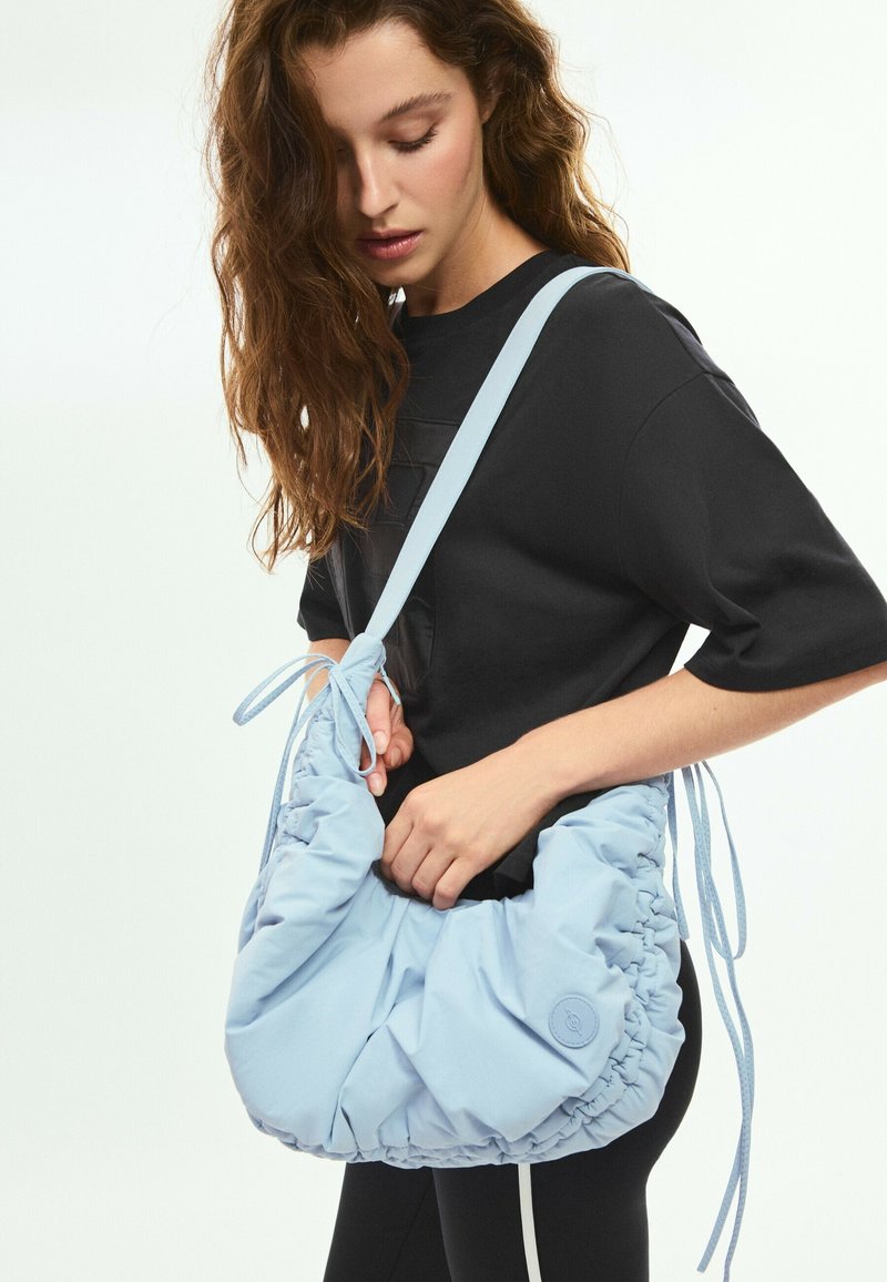 Jeune femme aux longs cheveux bouclés portant une chemise noire tient un grand sac à épaule bleu clair froncé avec des bretelles réglables.