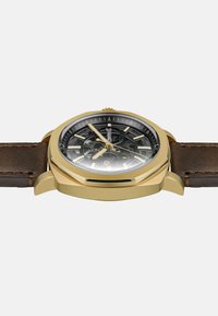 Orologio da polso in tonalità oro con quadrante scheletrato nero, indicatori dorati e cinturino in pelle marrone, mostrato di lato su sfondo bianco.