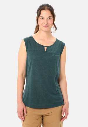 Vrouw draagt een mouwloze donkergroene top met een kleine sleutelgatopening aan de voorkant en beige broek, staand tegen een effen lichte achtergrond.