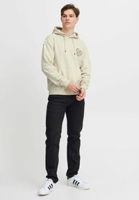 Felpa con cappuccio beige, dotata di tasca a canguro e patch rotondo con logo, abbinata a jeans neri e sneakers bianche con strisce nere.