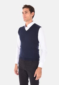 Maglione senza maniche di colore blu navy, design a scollo a V, realizzato in materiale lavorato a maglia, abbinato a una camicia bianca con colletto e pantaloni neri.