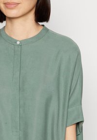 Zelený textilní top s kulatým výstřihem, předním zapínáním na knoflíky a krátkými rukávy. Materiál vypadá jemně s jemnou strukturou.