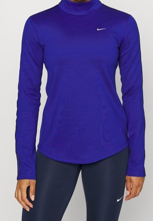 Žena nosící královsky modrý atletický top s dlouhým rukávem a tmavě modré legíny, obojí s malými bílými logy Nike.