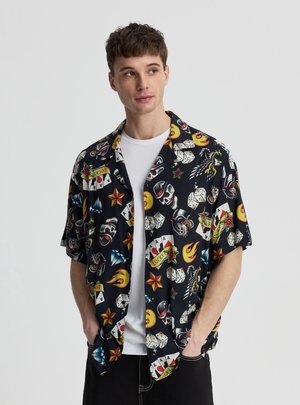 Młody mężczyzna w czarnej koszuli z krótkim rękawem z kolorowymi wzorami w stylu tatuażu na białej koszulce, ręce w kieszeniach, patrzy w prawo.