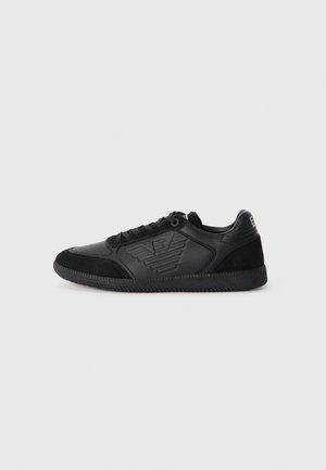 Black sneaker featuring a mix of smooth leather and suede, perforated toe, raised logo accents, and a textured rubber sole.