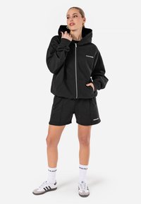 Čierna mikina na zips s prednými vreckami a logom "PEGADORE". Skombinovaná s čiernymi šortkami. Model má na sebe biele ponožky a čierne tenisky.