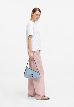 Geantă triunghiulară din denim, cu margini negre și logo-ul K, ținută de un model care poartă un tricou alb și pantaloni largi de culoare roz deschis.