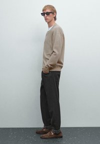 Maglione beige a maniche lunghe, pantaloni larghi grigio scuro e mocassini marroni, abbinati a occhiali da sole neri rettangolari.