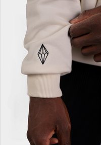 Felpa crema con polsini a costine e un ricamo nero a forma di diamante sul maniche sinistra; texture soffice, design casual.