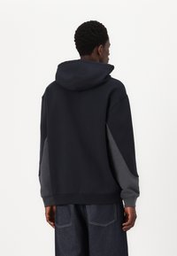 Sweatshirt à capuche bleu marine avec des manches grises contrastantes, coupe ample, texture douce, poignets et ourlet côtelés. Ne comporte aucun logo ni motif visible.