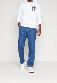 Hoodie bianca con tasca a marsupio e patch con logo, abbinata a jeans blu a taglio rilassato e sneakers bianche. Tessuto morbido, design casual.