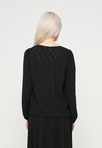 Γυναίκα με μακριά πλάτινου ξανθά μαλλιά, φορώντας μια μαύρη μακρυμάνικη υφασμάτινη μπλούζα και μαύρη φούστα, απεικονίζεται από πίσω σε απλό φόντο.