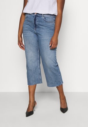 Frau trägt hellblaue, verkürzte, weit geschnittene Jeans mit rohem Saum, weißes Lochmuster-Top und schwarze spitze High Heels, steht auf grauem Boden.