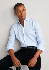 Sjedeći muškarac u svijetloplavoj košulji s dugim rukavima podvrnutim i crnim hlačama s crnim remenom, gleda u stranu.