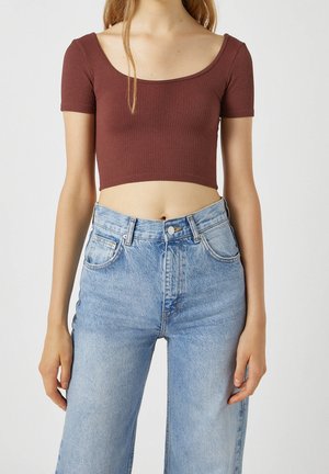 Femme portant un top court marron à manches courtes et un jean en denim bleu clair taille haute sur un fond uni.