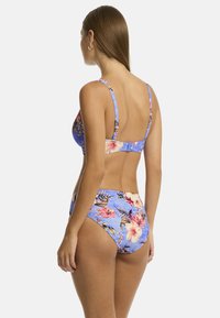 Modella con capelli castani lisci che indossa un bikini blu con un motivo floreale, rivolta di spalle contro uno sfondo bianco puro.