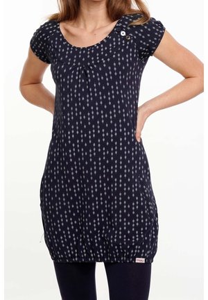 Nainen käyttää tummansinistä lyhythihaista tunikaa, jossa on valkoinen kukkakuvio, pyöreä pääntie, napit olkapäällä ja laskostettu helma mustien legginsien päällä.