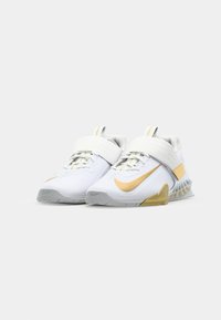 Zapatillas deportivas blancas con acentos dorados, que cuentan con una amplia correa de velcro, material superior texturizado y un diseño distintivo de suela ondulada.