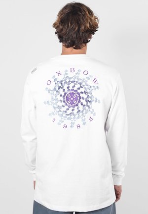 Homme portant une chemise blanche à manches longues avec un motif en spirale géométrique violet et bleu et des lettres au dos, dos à la caméra.