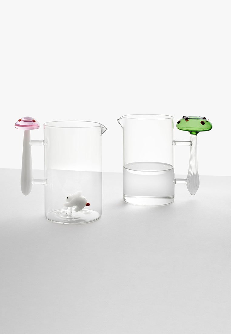 Dwa przezroczyste dzbany szklane z unikalnymi uchwytami: jeden w kształcie różowego grzyba z białym zwierzęciem w środku, drugi z zielonym uchwytem w kształcie grzyba.