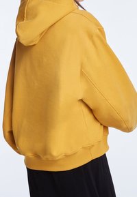 Sudadera amarilla con bolsillo tipo canguro, puños acanalados y capucha con cordón, que presenta una textura suave y un ajuste relajado.