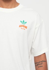Hvid bomulds T-shirt med rund halsudskæring, forsynet med et teal tre-blads logo og orange tekst, der siger "ADIDAS" over tre flammer.