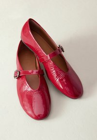 Red patent leather Mary Jane flats with a rounded toe, adjustable buckle strap, and smooth, glossy texture. Luxe sheen finish.