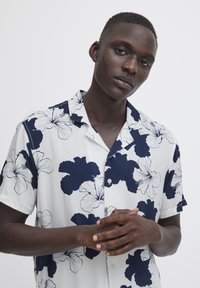 Camicia a maniche corte con bottoni di colore bianco con motivo floreale blu navy. Presenta un colletto rilassato e dettagli con bottoni. Tessuto morbido al tatto.