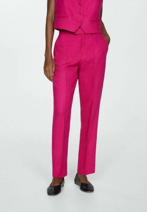 Femme portant un pantalon rose vif sur mesure et un gilet sans manches assorti, associé à des chaussures plates noires, debout devant un fond uni.