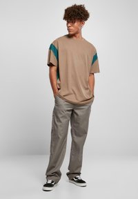 Beige Kurzarm-T-Shirt mit türkisfarbenen Akzenten, kombiniert mit hellgrauen Hosen und schwarzen Sneakern. Das Outfit hat eine entspannte Passform und eine weiche Textur.