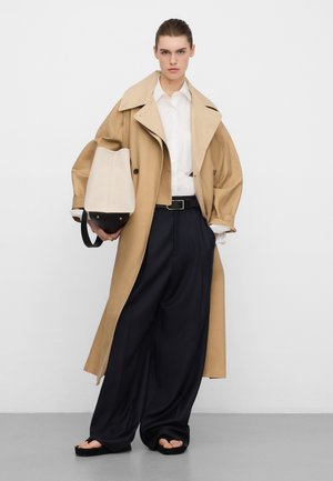 Beiger Trenchcoat über einem weißen Hemd, kombiniert mit schwarzen, weit geschnittenen Hosen. Hält eine beige-schwarze Handtasche mit glatter Textur.