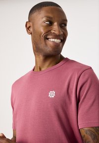 Camiseta de manga corta y cuello redondo en algodón rosa suave con un estampado blanco del logo en el lado izquierdo del pecho. Textura suave, ajuste casual.