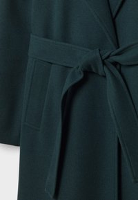 Manteau en laine vert foncé avec une ceinture en tissu nouée à la taille et une poche rectangulaire patch sur le côté.