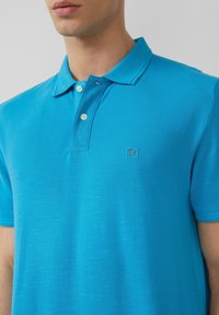 Türkises Polohemd aus strukturiertem Stoff. Verfügt über einen Polokragen, zwei Knöpfe und ein kleines, auf der Brust gesticktes Logo.