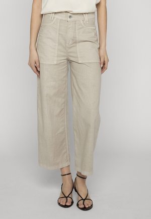 Kvinne iført beige vidbukser med frontlommer og svarte sandaletter med hæl, stående mot en enkel lys bakgrunn.