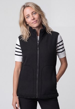 Blonde woman wearing a black zip-up fleece vest over a white short-sleeve shirt with black stripes on the sleeves, standing against a plain background.