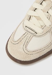 Bézs sportcipő, velúr és vászon anyagokkal, vastag krémszínű fűzőkkel, barna gumi talppal és kontrasztos fekete varrási részletekkel.