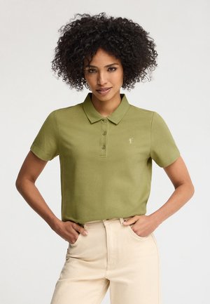 Kobieta o kręconych włosach, ubrana w oliwkowozieloną koszulkę polo z krótkim rękawem i beżowe spodnie, z rękami spoczywającymi w przednich kieszeniach.