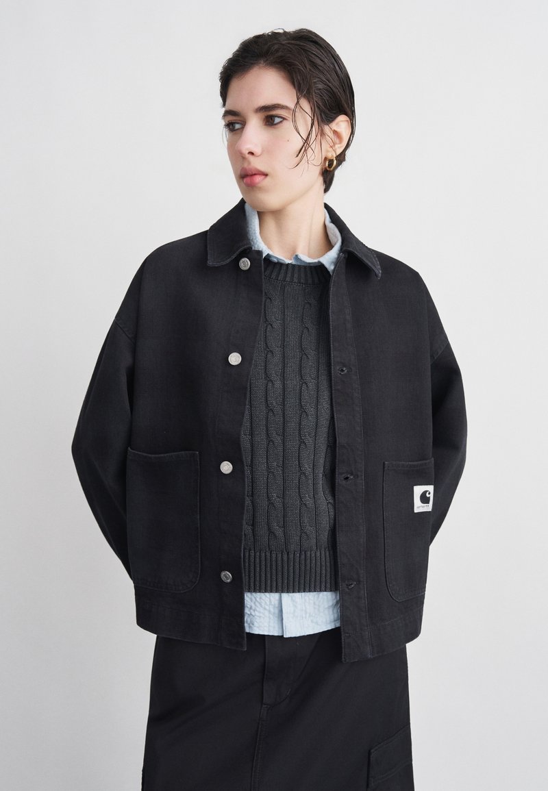 Person wearing black button-up jacket over dark gray cable knit sweater and light blue collared shirt, looking slightly to the side.