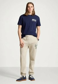 Giovane uomo con capelli lunghi fino alle spalle, indossa una t-shirt navy di Polo Ralph Lauren, pantaloni sportivi beige con coulisse e sneakers navy, in un ambiente neutro.